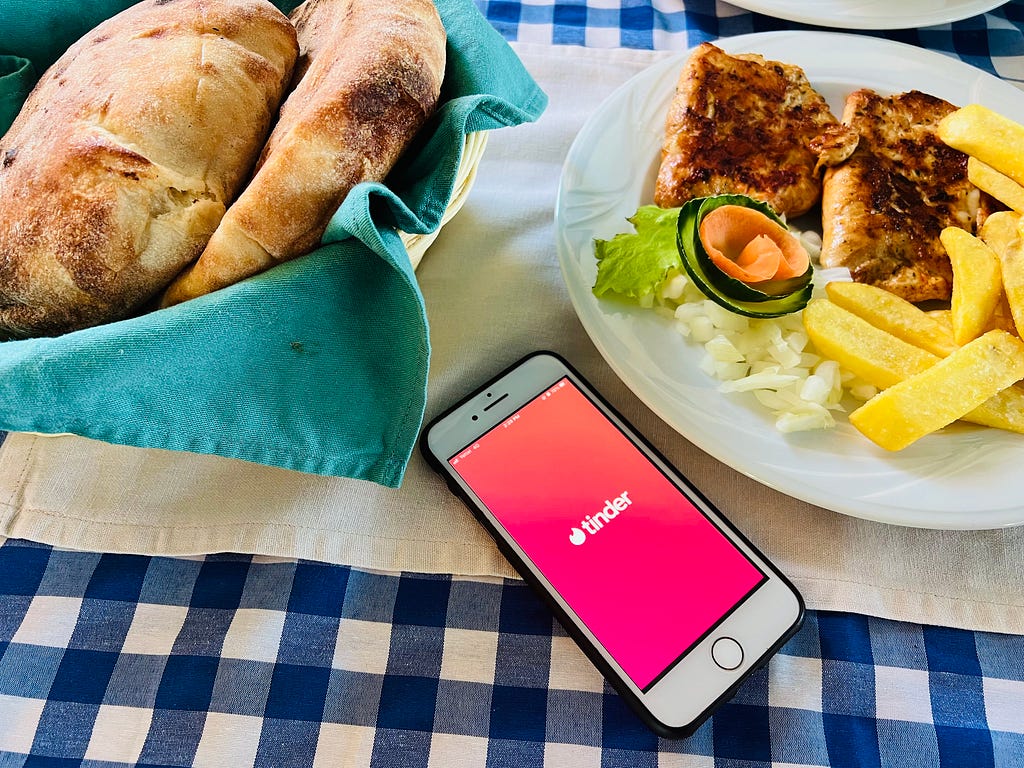 A phone with the Tinder app, sitting on a blue checkered tablecloth and surrounded by traditional Serbian food such as bread and meat and fries.