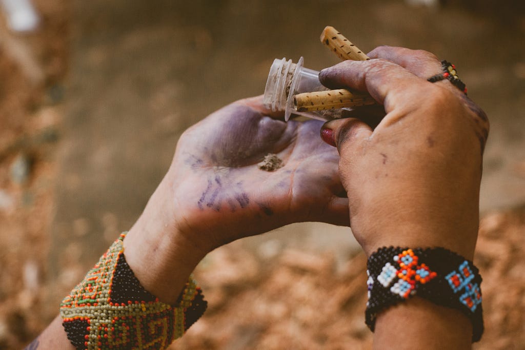 The hands of a person, with beaded bracelets encircling their wrists, hold a small canister of ancestral medicine and dispense it into their palm.