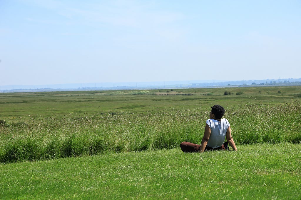 A guest sits on green grass. They wear a white top and red trousers. There is a blue sky above and in the distance. The face away from the camera with the hands behind them, holding them up as they look ahead.