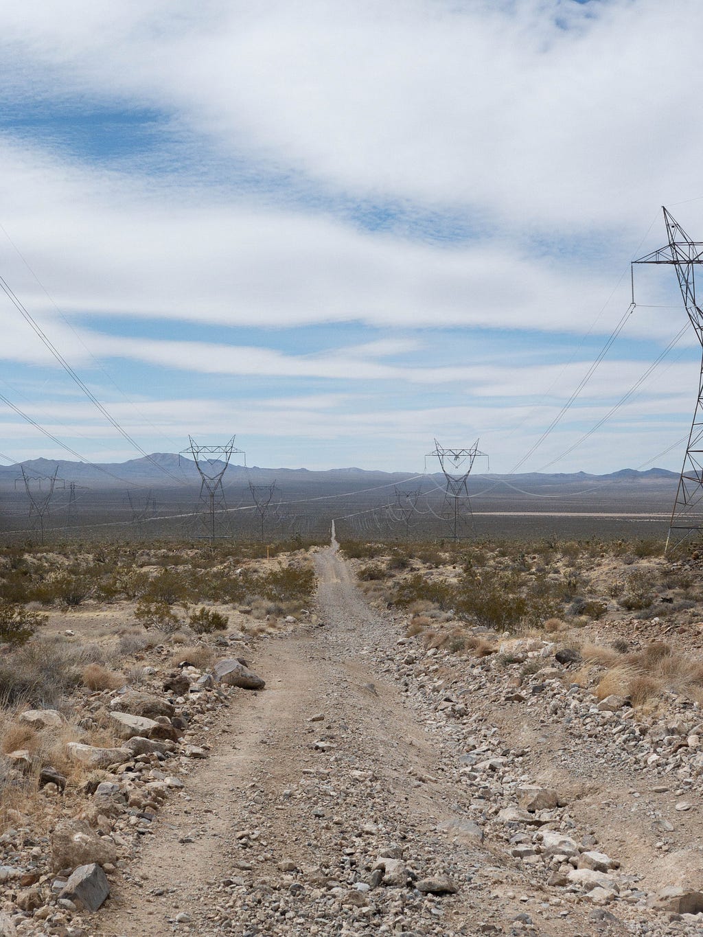 Looking backwards at a rocky and uneven dirt road going up a mountain. The road stretches into the desert. Road has long distance transmission towers on either side