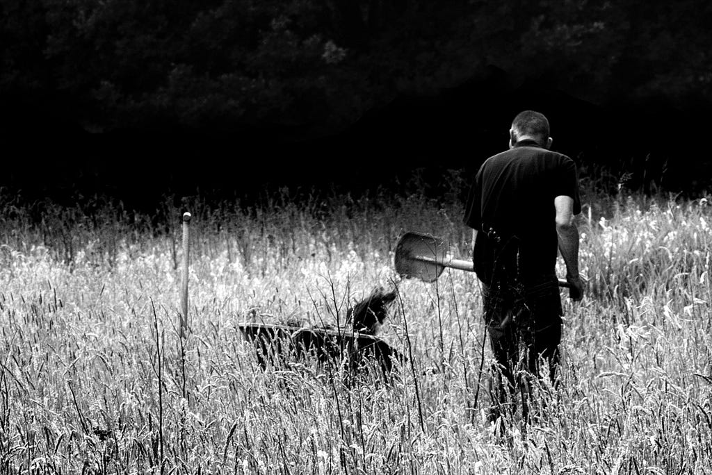 Man wearing black standing in a field facing away from camera. He is holding a shovel, tipping dirt into a wheelbarrow.
