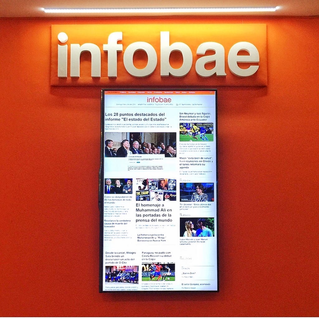 A computer monitor displays the homepage of infobae.com, an Argentine news website. The wall behind it is bright orange.