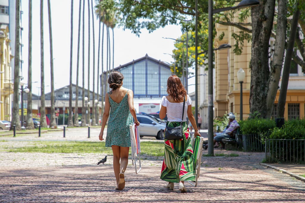 Duas mulheres, de costas, caminham lado a lado na Praça da Alfândega segurando cadeiras de praia