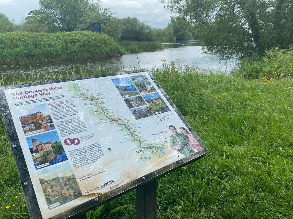 A sign shows the route of the Derwent Valley Heritage Way. In the background are a canal and river that merge together.