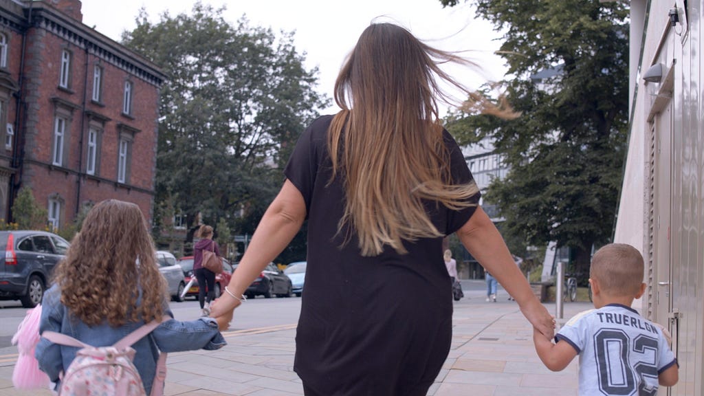 A woman with long blonde hair who is wearing a black t-shirt walks away from the camera holding the hands of two young children.