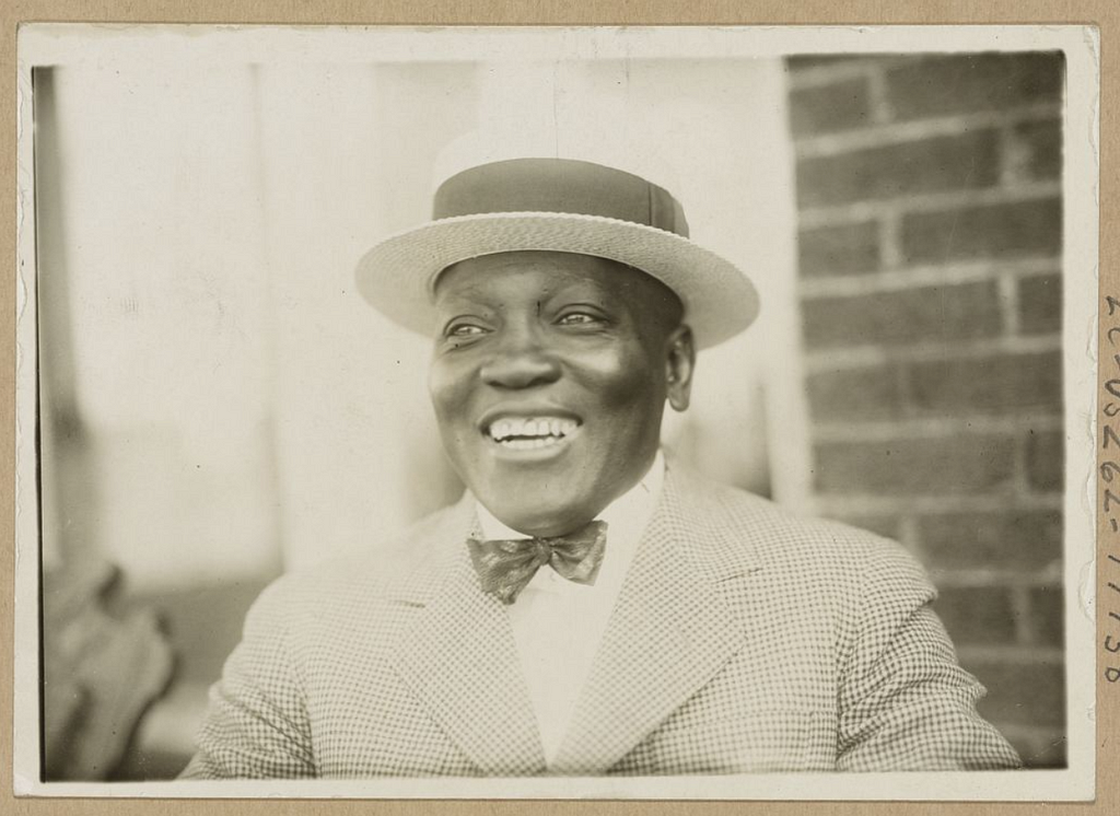 A black and white photograph featuring a smiling man with brown skin and wearing a hat, jacket, and bowtie