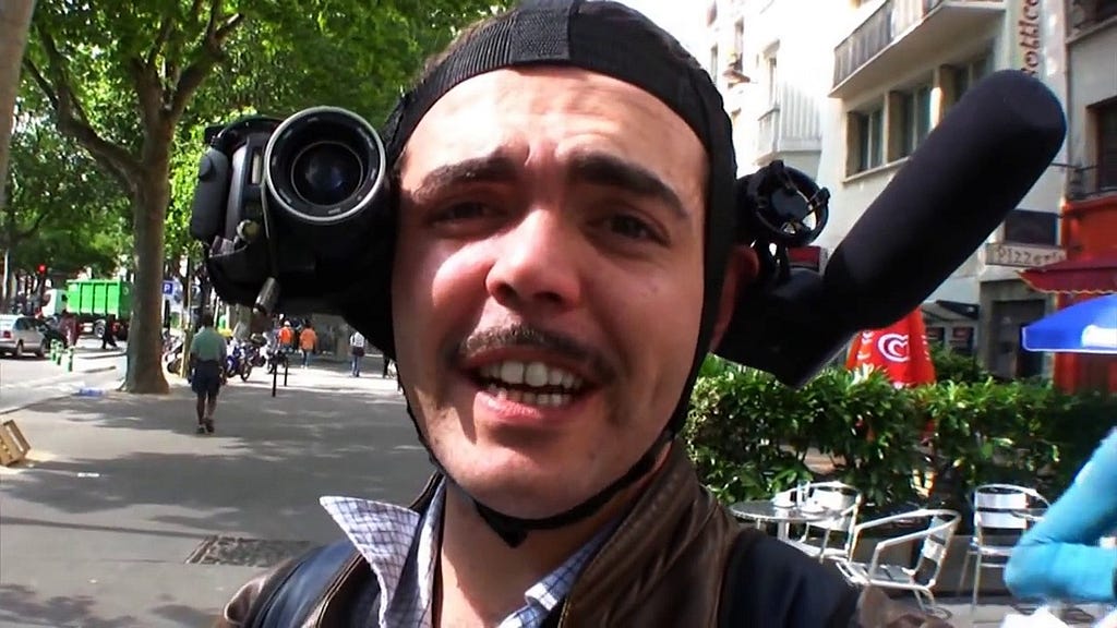 A mustache wearing man with a weird contraption around his head is seen smiling at the camera.