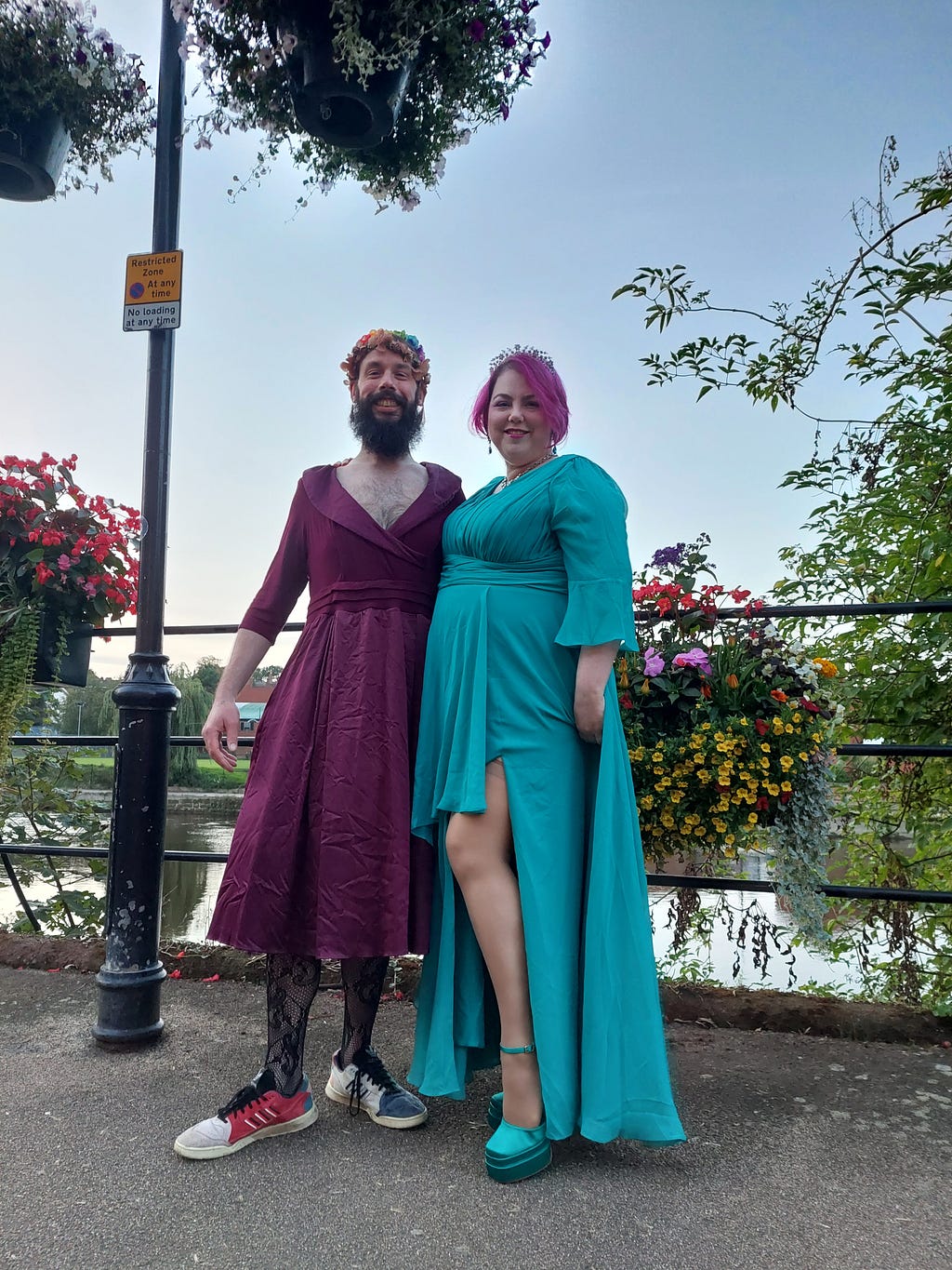 Two people wearing ballgowns, stood with containers of flowers by a river. The person on the Left presents as male, 6 foot 3 inches, with dark hair and a long beard. They are wearing a flower crown, fishnet tights and trainers. The ballgown is burgundy satin, reaches to mid-calf and has a tuxedo-style bodice and a full skirt over net. Claire, right, is standing in a floor-length, empire-line, teal chiffon ballgown with a thigh-high split, 7 inch platform heels & a diamante tiara on her pink hair