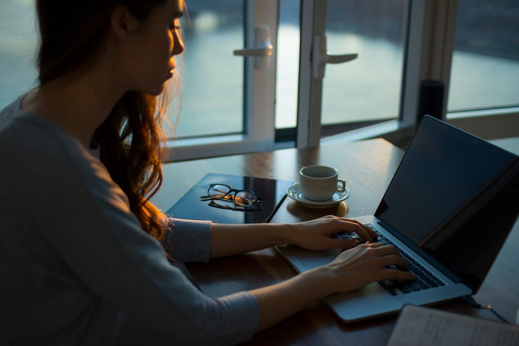 A woman with long, dark hair sits at a desk with a laptop and coffee.