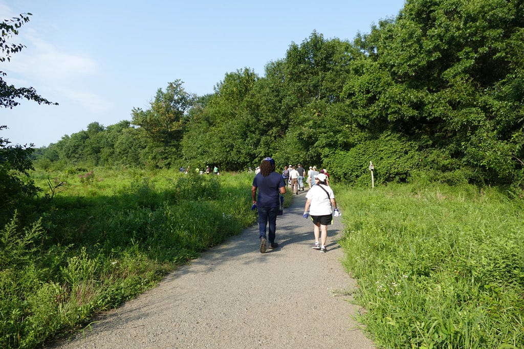 A group of about 20 people distanced a few feet apart walk along a trail that passes through green fields and along forest.