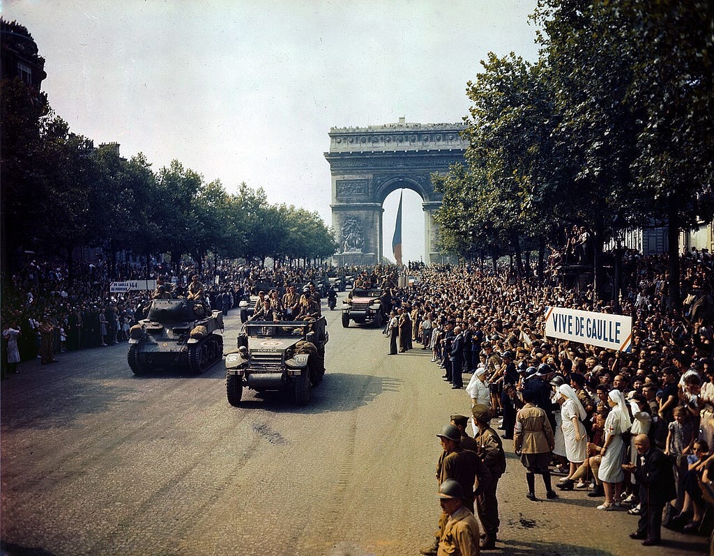 Des véhicules de la 2e division blindée descendent les Champs-Élysées entre une foule dense, sous l’Arc de Triomphe, le 26 août 1944.
