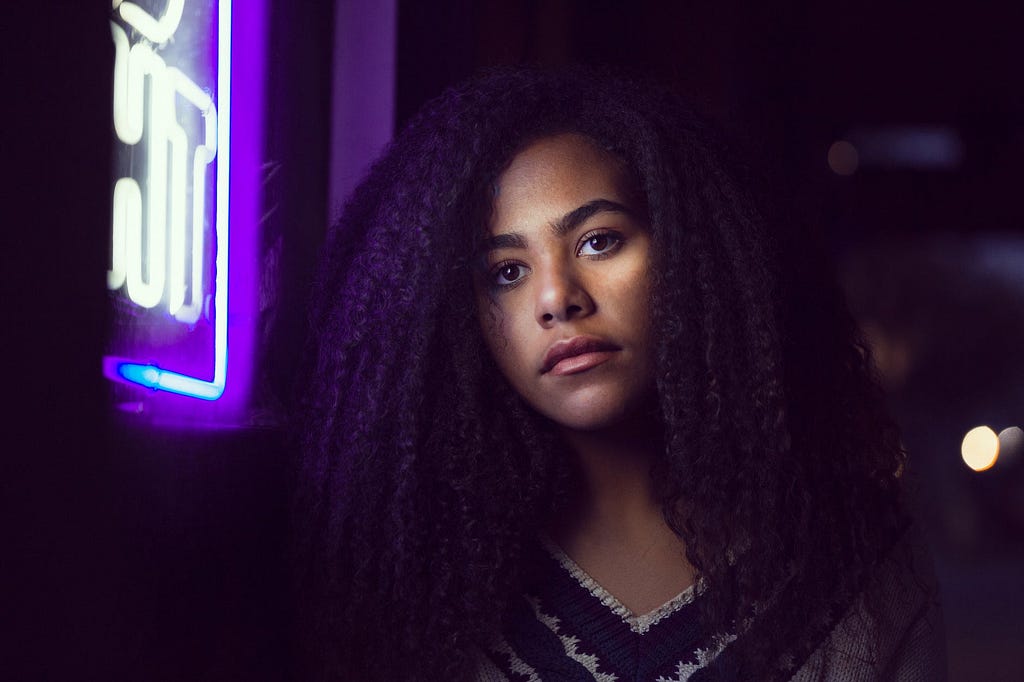 A woman standing next to a neon sigh looks into the camera