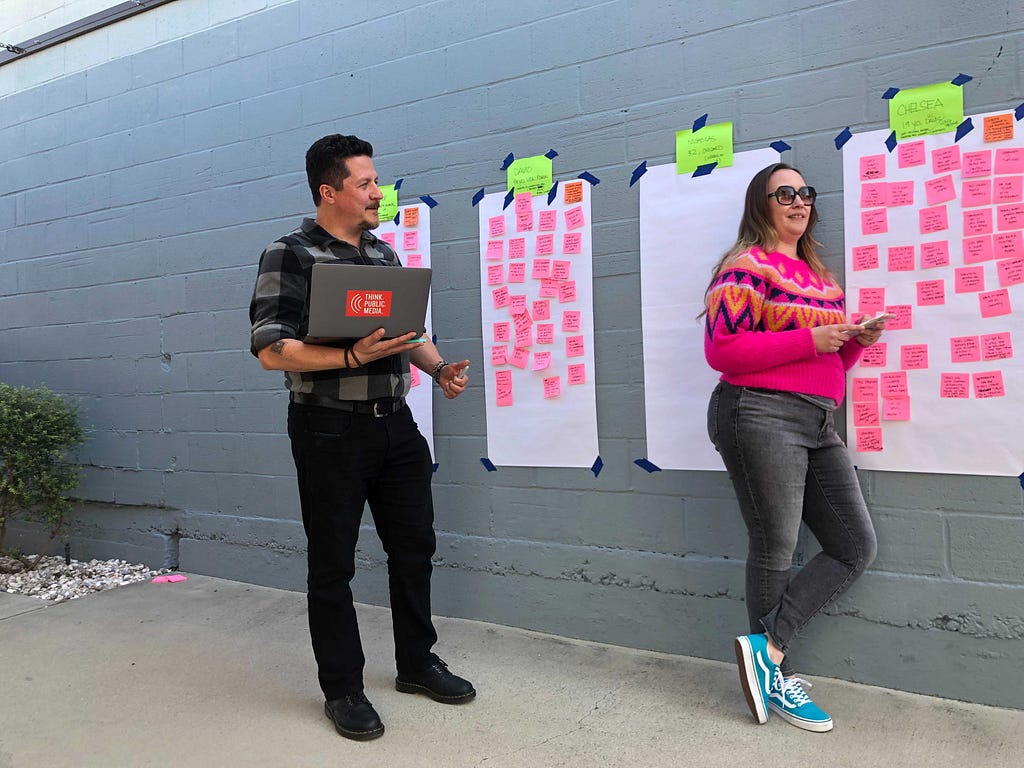 A man and a woman stand casually in front of a gray wall with oversize Post-its populated with several bright pink Post-its. The man wears dark clothing and holds a laptop. The woman, who is to the right and leaning against the wall, wears bright blue shoes, dark jeans, and a pink sweater with glasses.