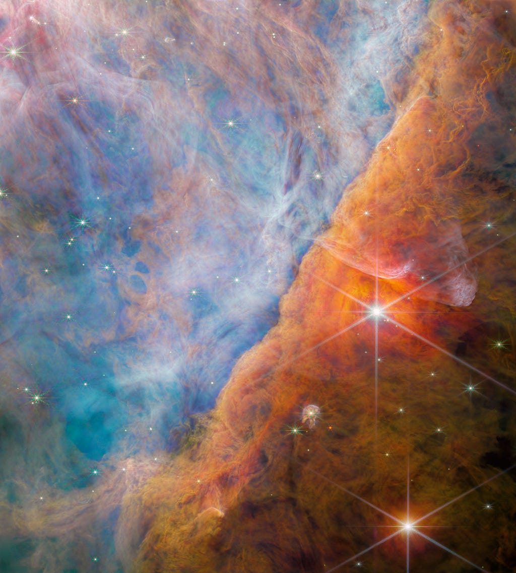 An image by the James Webb Space Telescope of a section of the Orion Nebula. Billowy, multi-hued clouds fill the field of view. The scene is divided by an undulating formation running diagonally from lower left to upper right. On the left side, the clouds are various shades of blue with some translucent orange wisps throughout. On the right side, the clouds vary from bright orange-red to brown as you go from left to right.