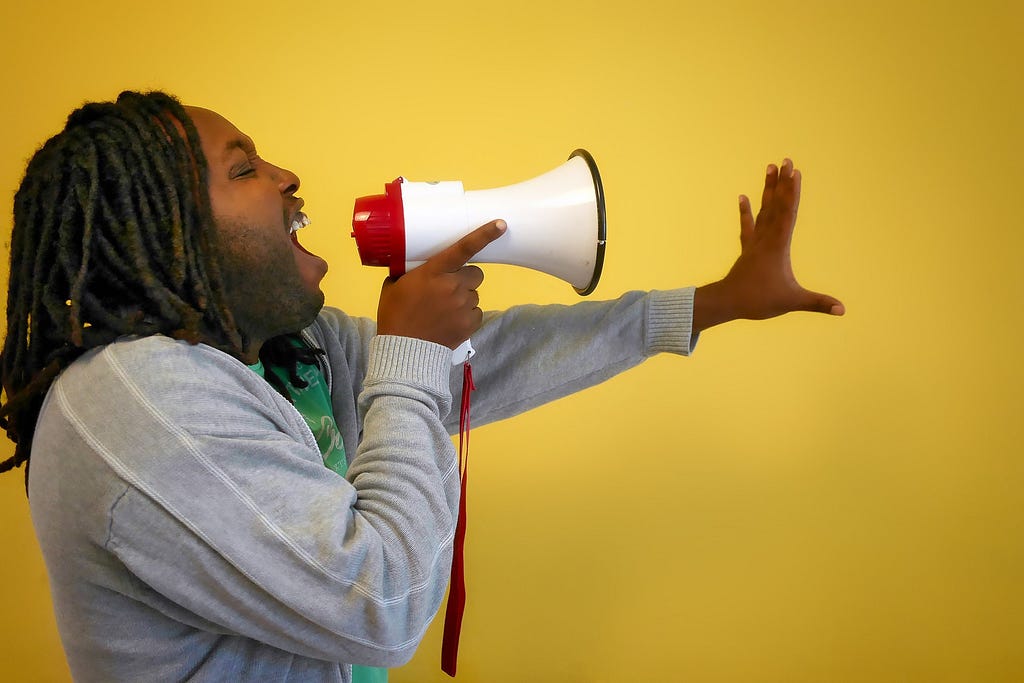 a dark brown skinned person with shoulder length dreadlocks shouting into a megaphone with their hand outstretched in front