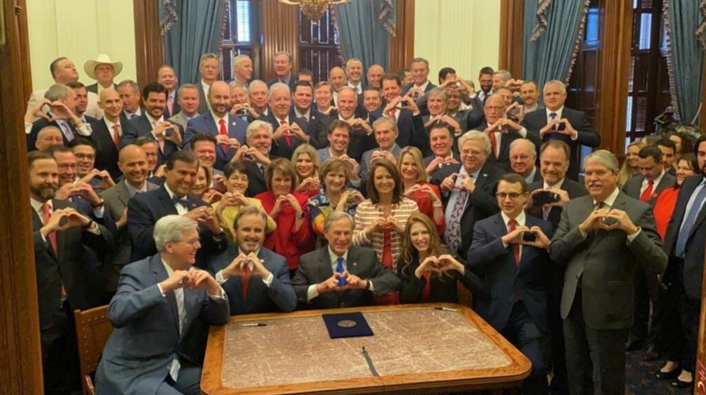 72 Texas legislators holding up their hands to make a heart shape as they stand behind Governor Abbot and the recently signed bill