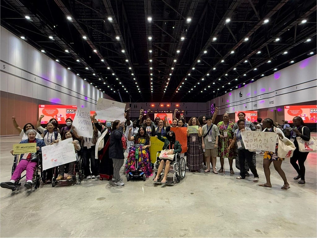 Un grupo de mujeres, algunas en sillas de ruedas, levantan su puño o pancartas con consignas feministas. Están en un lugar cerrado dentro del Foro AWID.