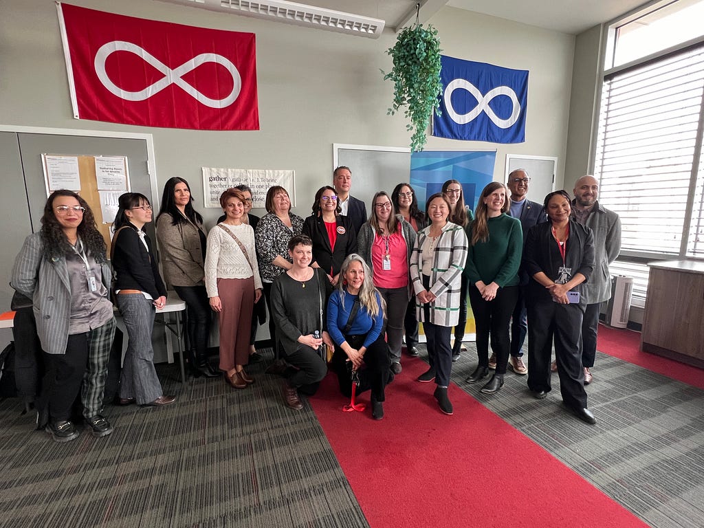 A group of housing providers, City Councillors, and the Mayor pose infront of Metis flags at a media event in a large community room.