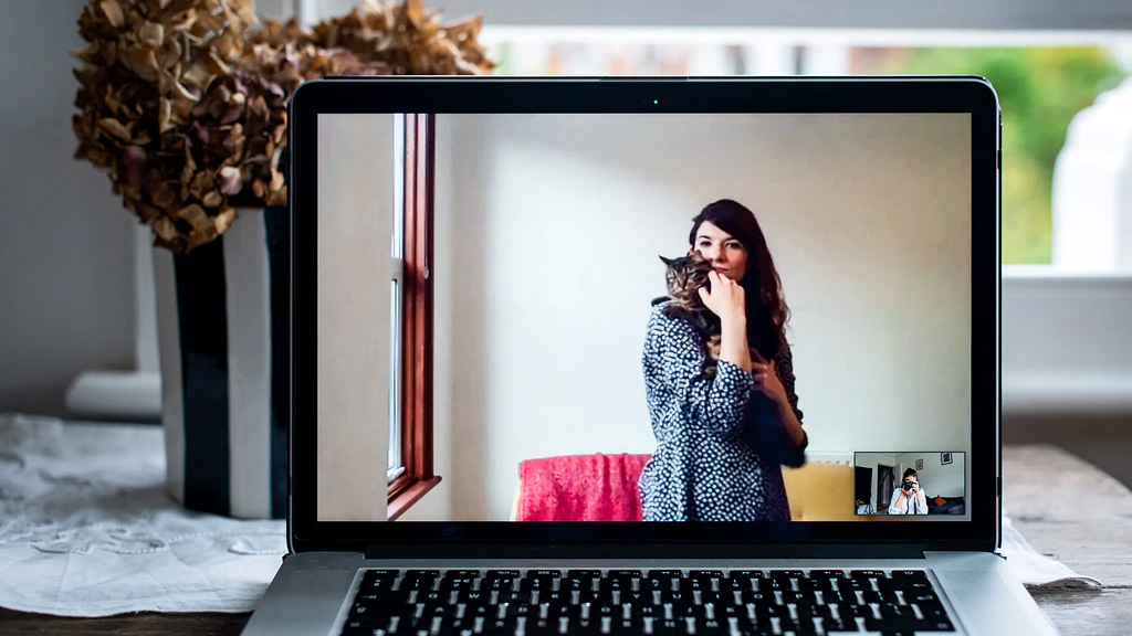 Photograph of an open laptop within a domestic scene. Most of the image is taken up with the screen, with part of the keyboard and trackpad visible. On the screen is a video call showing a woman standing in her living room, holding a cat in her arms, both are looking to camera. In the bottom right corner of the screen the photographer can be seen in a small floating window, camera to her eye, in the process of taking the picture.