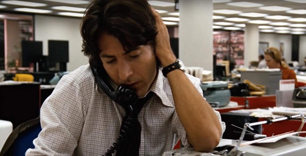 Still from the movie All the President’s Men. Dustin Hoffman on the telephone in a newsroom with his hand over his ear.