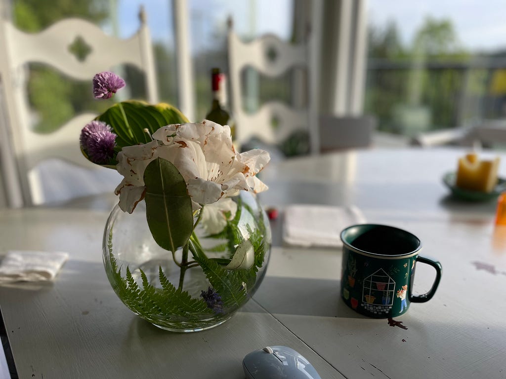 A white and purple flower arrangement in a round clear vase, in the morning light on a table next to a coffee mug. A spent wine bottle in the background.