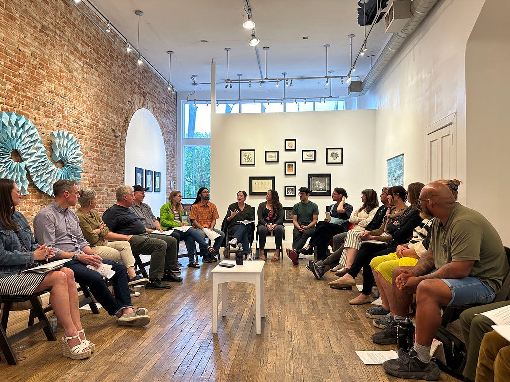 A group of 30 people sits in a circle in a room with a one red brick and two white walls. They are engaged in conversation. A white table with a tape recorder rests at the center. On the walls are various works of art.