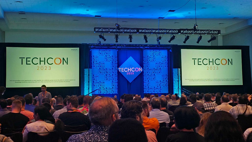 A conference room full of people looking at a stage that displays the TechCon 2023 logo.