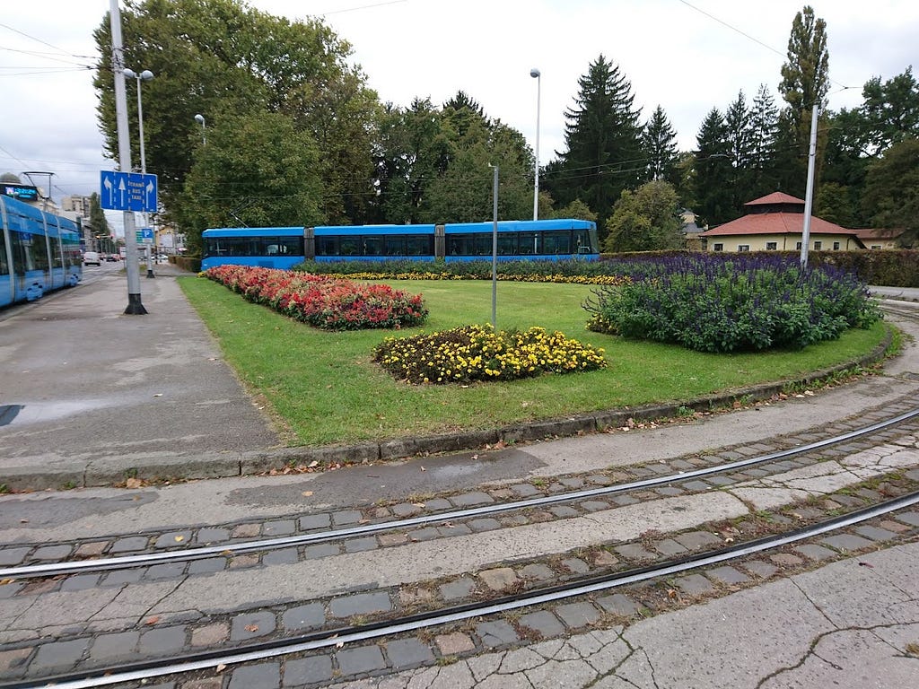 A blue tram is parked next to an empty sidewalk by a spacious green park entrance