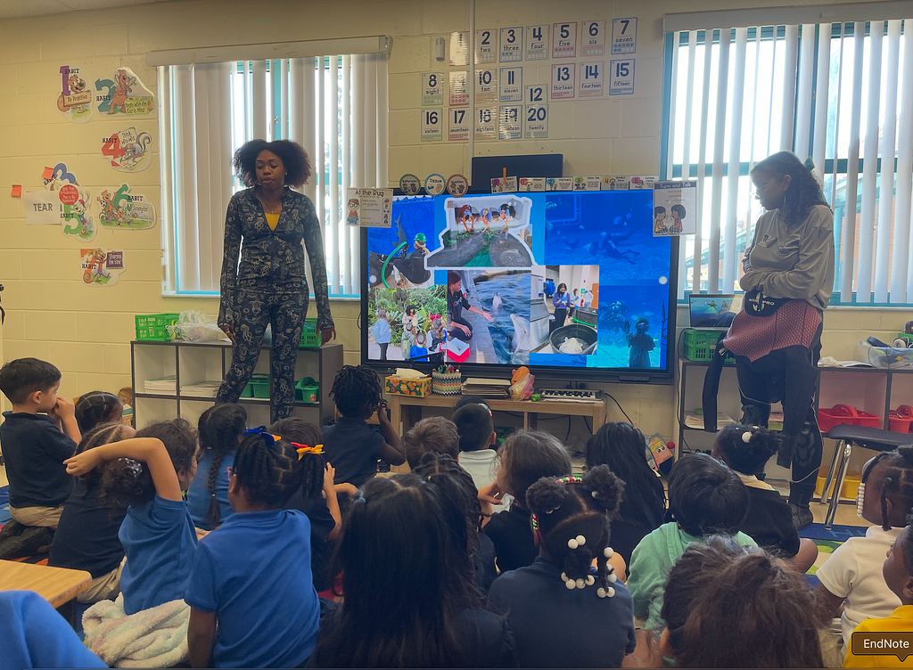 A classroom filled with young students seated on the floor, facing the front of the room. Raven and MJ stand near a large television screen displaying a collage of photos related to marine life, fieldwork, and conservation activities. MJ is wearing a patterned long-sleeve outfit, and Raven is wearing a light-colored top layered with a jacket and dark pants. The classroom walls are decorated with educational posters, numbers, and children&rsquo;s artwork, and large windows with blinds.