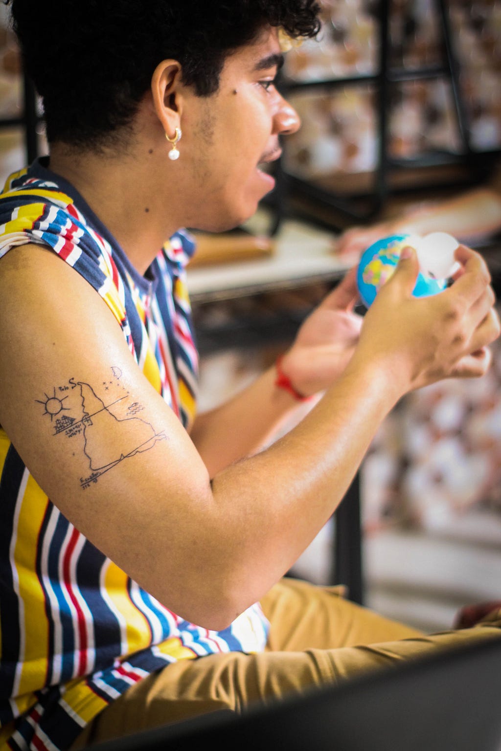 Jovem com explicando algo e segurando um globo terrestre nas maos. Tatuagem da America do Sul no braço direito.