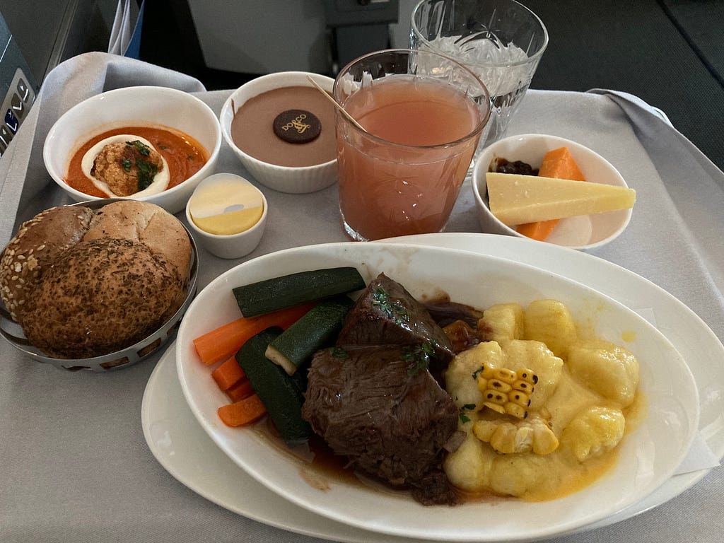 A photo of my dinner from my first time flying business class. Including a roll with three types of bread, a tomato dip, chocolate pudding, a summery cocktail, a cheese selection and the main event: a slow cooked beef rib and cheek with vegetables and sweetcorn gnocci.