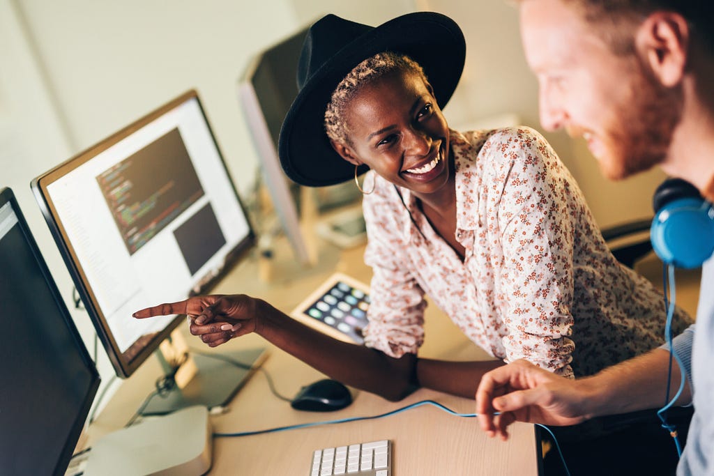 Two people sitting in front of a computer monitor with one person pointing and smiling and the other looking and smiling.