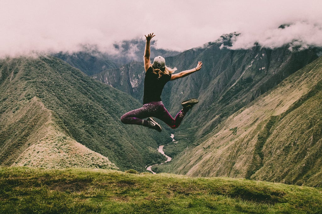 A woman jumps for joy. There is a view of mountains in front of her.