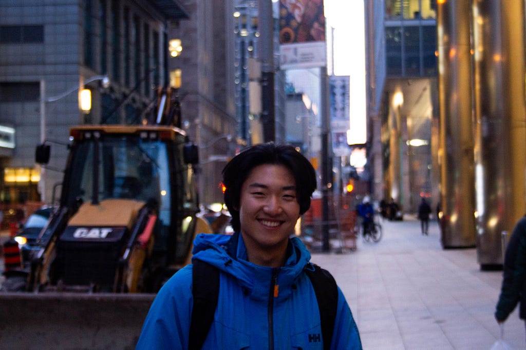 A profile photo of John. He is wearing a blue windbreaker, smiling, and standing on a city street with skyscrapers, a sidewalk, and construction machinery behind him.