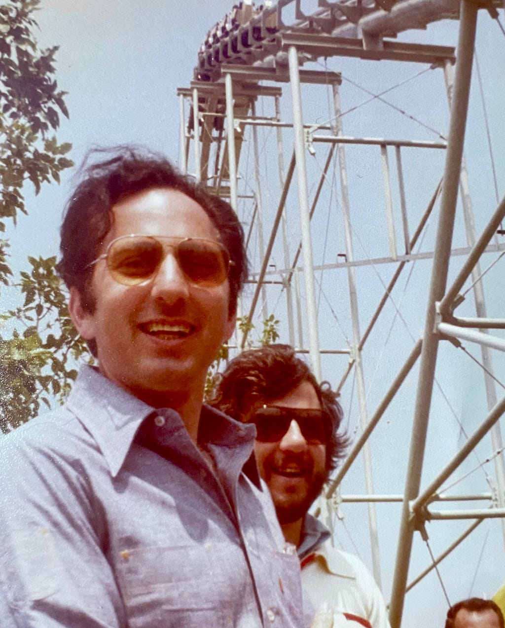 Two men with dark hair, both in sunglasses, in front of a steel structure smiling.