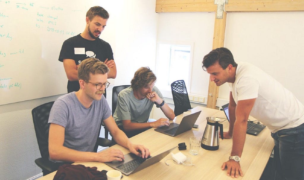 A group of young men sitting and standing around a table with their laptops. A whiteboard on the left with scribbles on it.