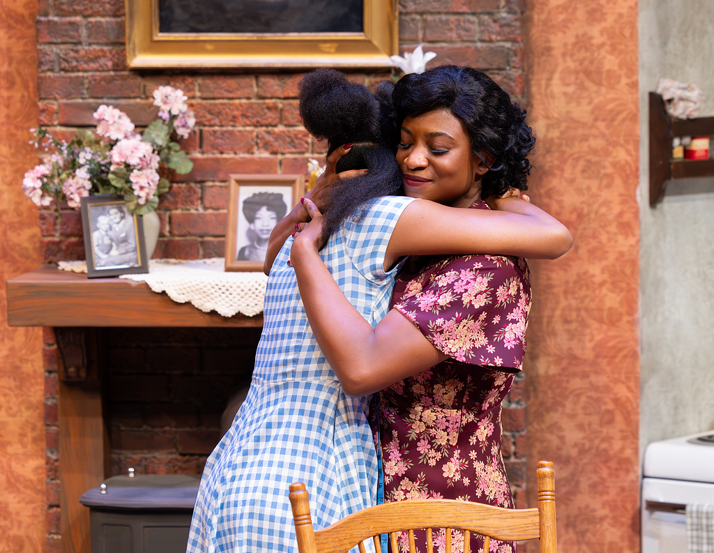 Two women in 1950s-era dresses hug near a brick wall in an apartment set.