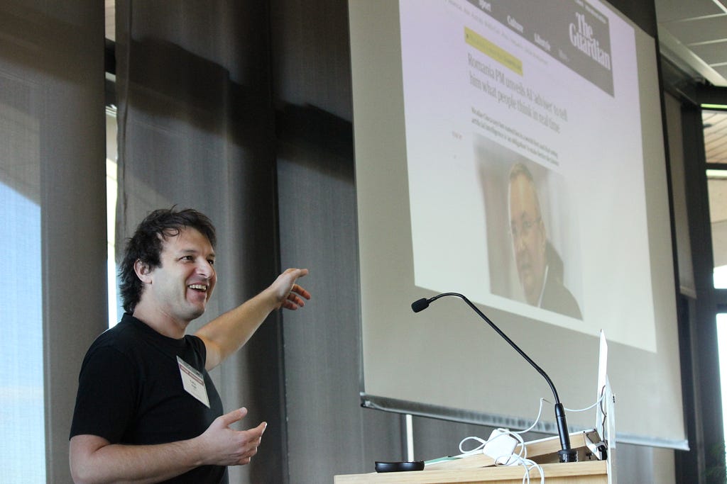 Colin Megill standing at a podium and pointing at a screen with a news article projected.