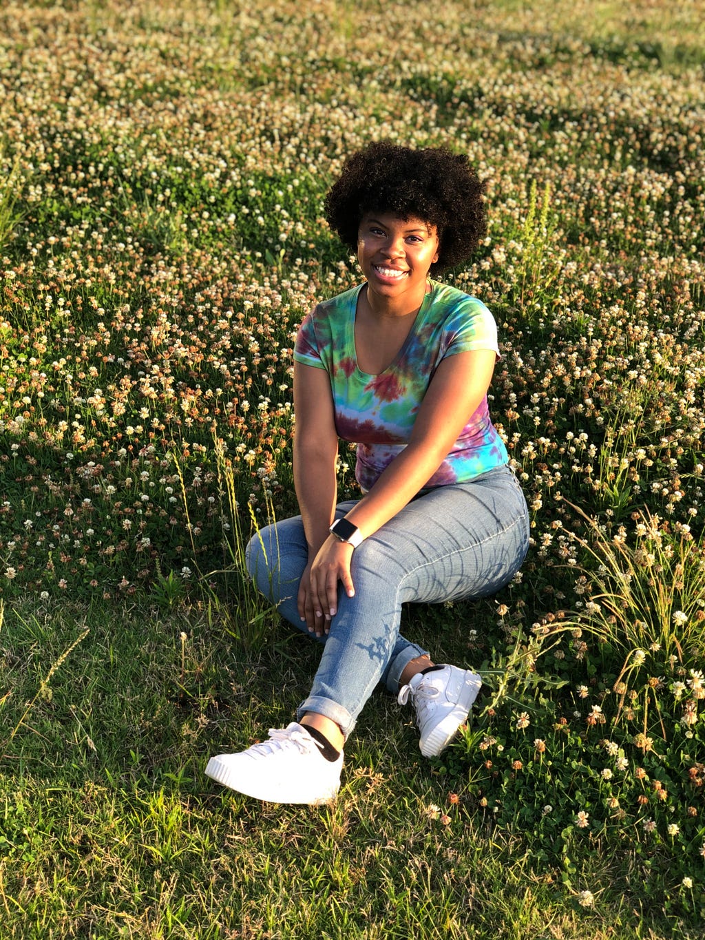 A black woman w/ an afro wearing a tie-dye t-shirt,blue jeans, and white sneakers sitting in a field.