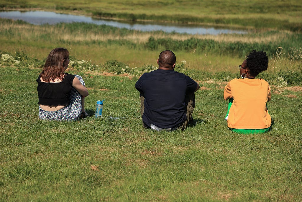 Three people (the Black Design Guild Care Team) sit on green grass facing away from the camera. On the left you have Amelia wearing a black top and black and white trousers. In the middle you have Michael wearing all black. On the right you have Tope wearing a orange top and green bottoms. Behind them is a small body of water.