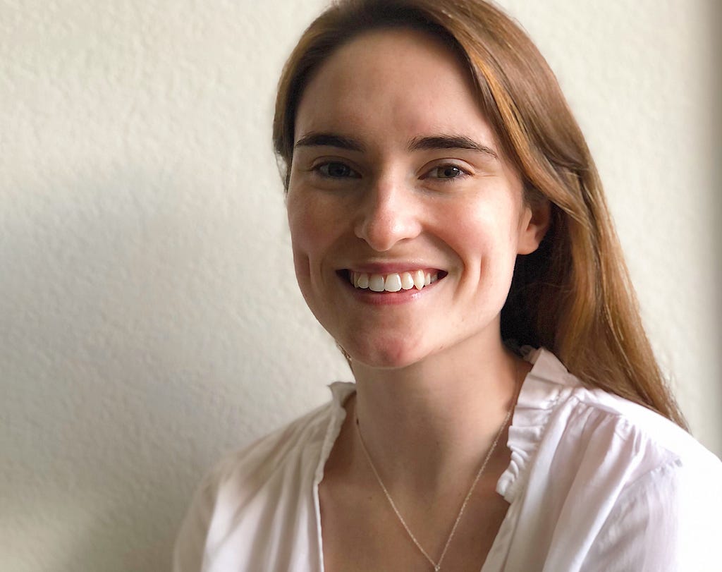 Marney, a young white woman with long reddish-brown hair draped down her back, is looking at the camera and smiling revealing dimpled cheeks. You can see her from her shoulders up and she is wearing a white blouse with a ruffled collar and a thin gold chain necklace.