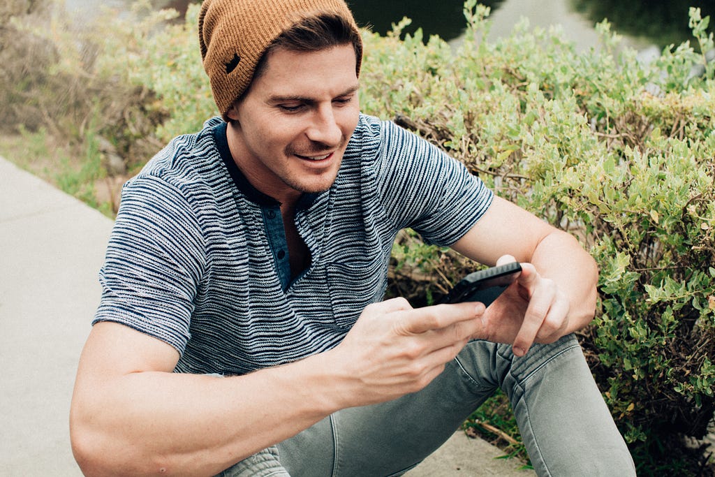 A man sits on a sidewalk smiling slightly as he stares at his phone