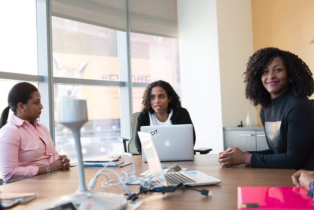 Three African American business women sitting at a brown, office table having a meeting with two laptops, electrical cables.