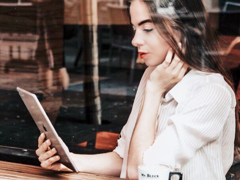 A woman sits at a bench, looking at her iPad. She is resting her left hand against her face.
