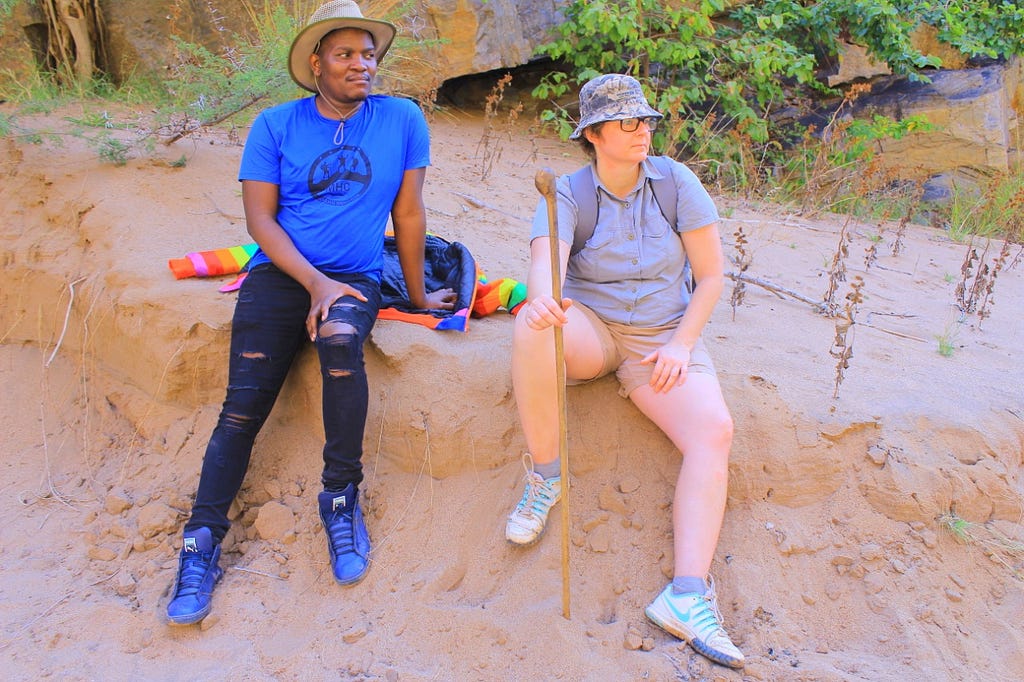 An African male and Caucasian female sitting on a sandy bank at the edge of a dry riverbed, looking in the same direction