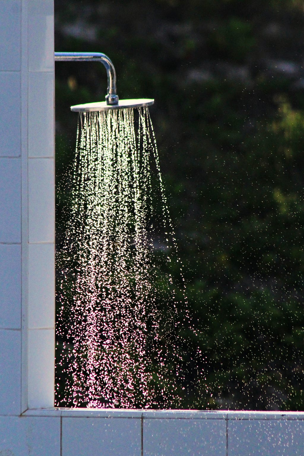 Photo of an outdoor shower, the rain-like showerhead is mounted on a white tile wall, as water flows out from it. Green nature is in the background.