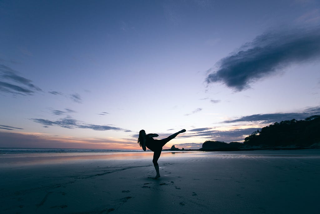 A person practices a high martial arts kick on a beach at sunset