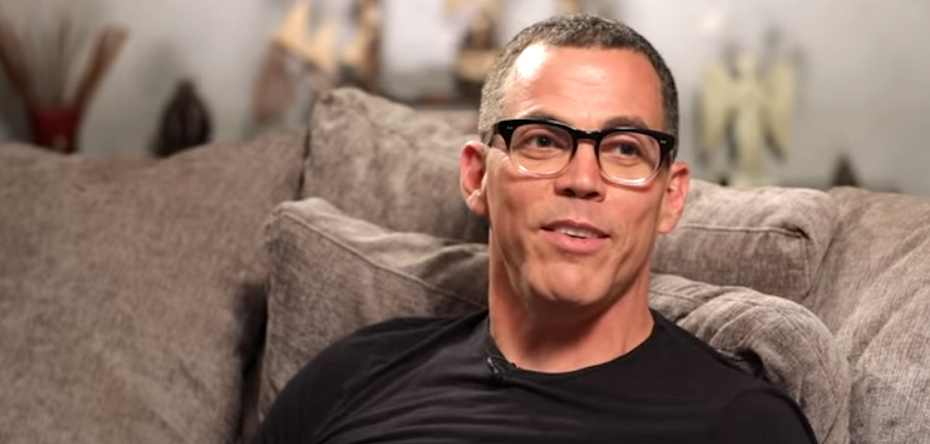 Steve-O, wearing glasses and a black shirt, sitting on an oatmeal colored couch in conversation with others offscreen