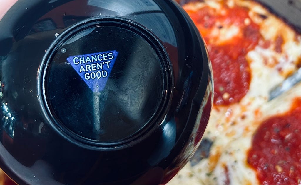A Magic 8-Ball toy displays the message, “Chances aren’t good.” In the background, a tantalizing pizza is topped with red sauce and pepperoni.