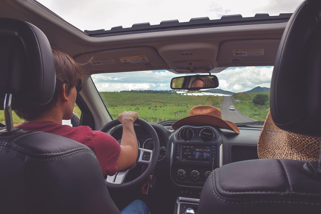 Man driving down a long road with a cowboy hat on the dashboard.